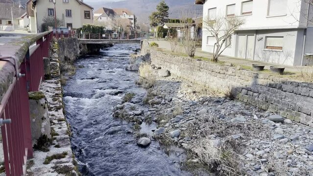 Small river flowing noisily among rocks and grass, under the road and an old concrete bridge with red barriers, in a village (Buhl, France), on a sunny winter day. Movement from bottom to top