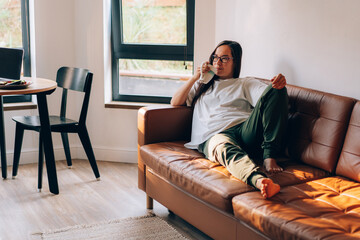 Fototapeta premium A young brunette in glasses and home clothes is sitting on a sofa in the living room drinking coffee