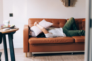 A stressed woman lies on the sofa in the living room and covers her face with a pillow.