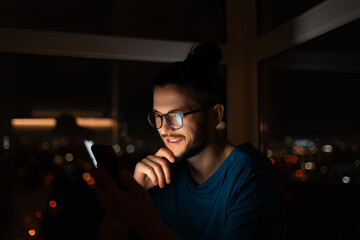 Indoor night portrait of young smiling man, holding smartphone.