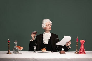 Portrait of young elegant man in peruke and vintage jacket sitting at table isolated on dark green...