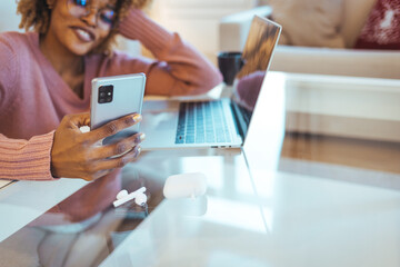 Business And Telecommuting Concept. Portrait of smiling young black woman using her smartphone and modern laptop computer, reading text message, sitting at desk with gadgets, working at home office