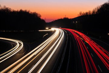 German Autobahn A46 at dusk on a winter evening in Iserlohn Sauerland at junction driveway town centre, long time exposure with light traces of passing car lights. Orange sky gradient after sunset. - obrazy, fototapety, plakaty