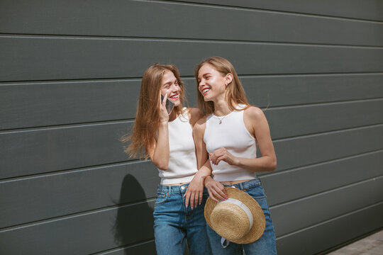 Generation Z twin sisters walking outdoors, laughing while one of them talking on phone.