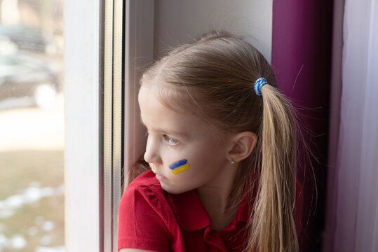 a crying child, looking out the window, flowing tears painted on her cheek in the yellow-blue colors of the Ukrainian flag, a request for help. Children ask for peace