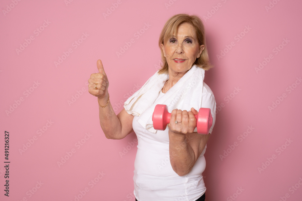 Wall mural smiling sport mature woman holding dumbbell showing thumb up isolated on pink background. active sen