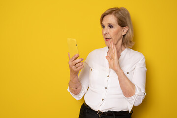 Happy mature senior woman using smartphone standing isolated over yellow background. Technology concept.