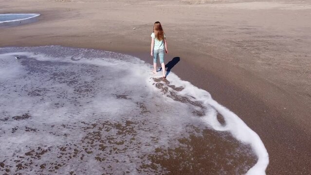 Girl with red shining hair walking on sandy coastline of California, USA