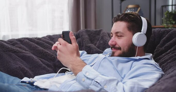 Portrait of adult man on sofa with smartphone in his hands listenng to music in headphones. Man chilling on couch. Male scrolling social media on gadget and nobing his head making dancing moves.
