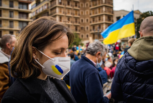 Upset Ukrainian Girl with Downcast Eyes Wearing a Mask Painted with the Colors of her Country and the National Flag in the Background in a Demonstration Against the War
