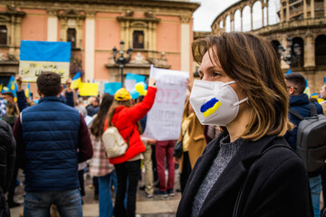 Ukrainian Woman with Determined Look Wearing Blue and Yellow Painted Mask in Demonstration in Favor of Ukraine and Peace With Protersters Behind - obrazy, fototapety, plakaty