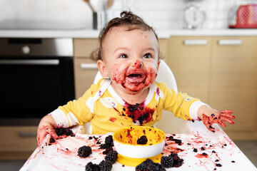 Funny baby with laughing face eating blackberries in high chair