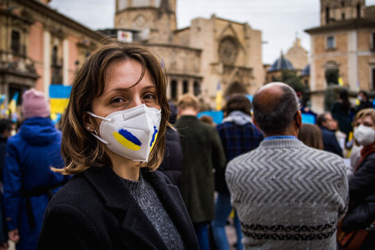 Ukrainian Girl Wearing Mask with Painted Ukraine Flag Blue and Yellow Looking at Camera with Demonstration Against War with Russia in the Background