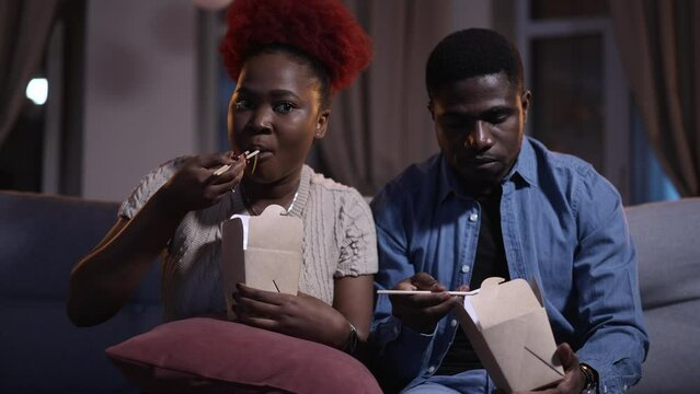 Two family people eating lunch and watching TV shows while sitting on couch at home spbas. 4k video Closeup view of young african american man, woman eat boxed food and watch movie with smiles, have