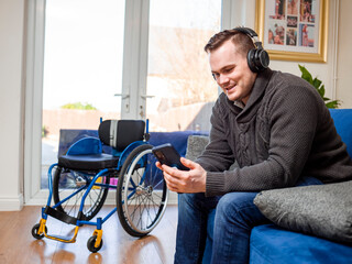 Man with headphones and smart phone sitting on sofa, wheelchair in background