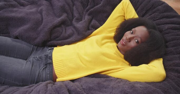Top view of african american wooman lying on sofa with arms crossed behing her head looking around. Female in yellow clothes. Tired female after work resting. Concept of rest, carefree.