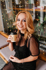 Young charming blonde with a cute smile and makeup while relaxing in a cafe. She is holding a cup of coffee in her hands. She is dressed in a black dress with transparent sleeves.