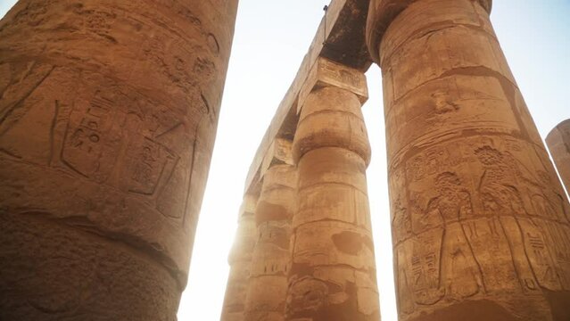 Massive pillars with intricate carved images and hieroglyphs in the Kamak Temple, Luxor, Egypt. 
Sun gleaming behind the pillars with a flare, slow sideways camera movement.