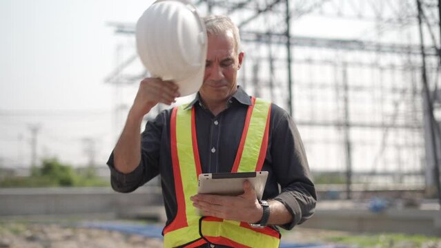 senior construction worker or architect using digital tablet checking structure in construction site outdoors with Hot weather sunny . builders taking off hard hat inspecting and working in workplace