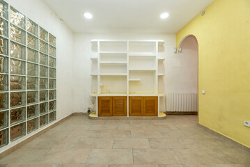 Empty room with plaster bookcase and low wooden furniture with Venetian style doors, round arch and glass pavé wall, yellow painted walls and gray terrazzo floor