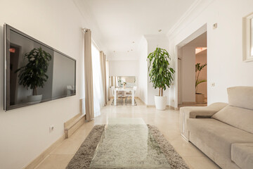 Long living room of an apartment with marble floors, cream sofas, tv on one wall and white dining table on the other side
