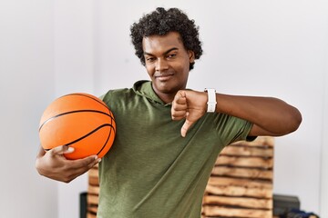 African man with curly hair holding basketball ball at the gym with angry face, negative sign showing dislike with thumbs down, rejection concept