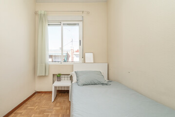 Single bed with blue bedspread with white headboard, matching side table and window with white curtains in a bedroom with parquet flooring