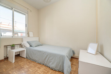 Single bed with blue bedspread with white headboard, matching side table and aluminum window with radiator below in bedroom with parquet floor
