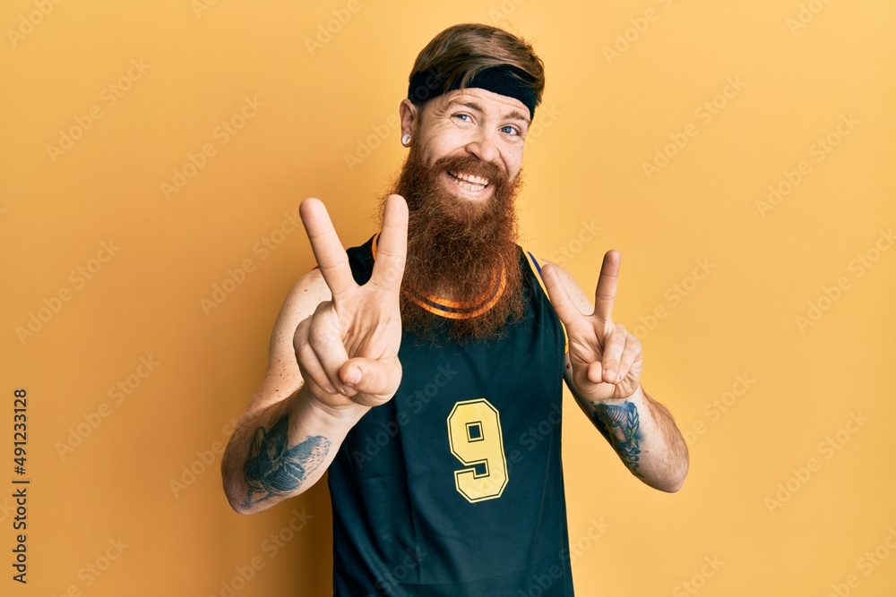 Sticker Redhead man with long beard wearing basketball uniform smiling looking to the camera showing fingers doing victory sign. number two.