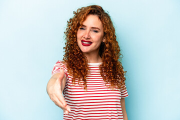 Young ginger caucasian woman isolated on blue background stretching hand at camera in greeting gesture.