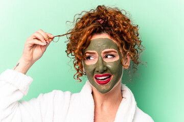 Young ginger woman wearing beauty facial mask isolated on green background