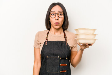 Young caucasian waitress woman holding tupperware isolated on white background shrugs shoulders and...