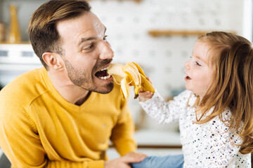 Happy little girl giving her father to eat banana in the kitchen