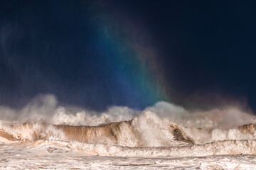 A close up view of a breaking wave with a small rainbow and a dark background