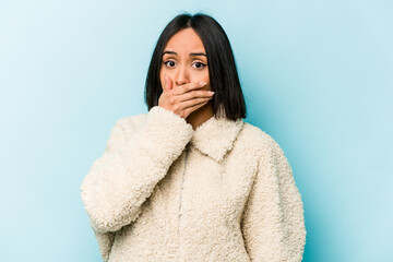 Young hispanic woman isolated on blue background covering mouth with hands looking worried.