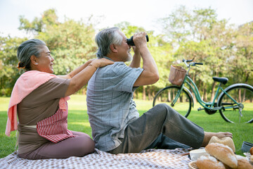 An elderly couple doing picnics and watching nature by binocular in the garden with bright smiles and enjoying the relaxation of summer season. Health and relaxation after retirement.