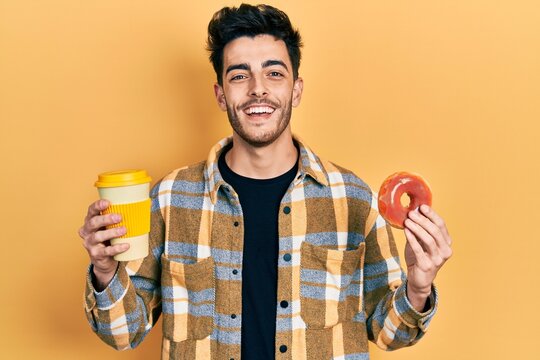 Young hispanic man eating doughnut and drinking coffee smiling and laughing hard out loud because funny crazy joke.