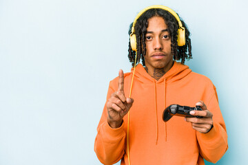Young African American man playing with a video game controller isolated on blue background showing number one with finger.