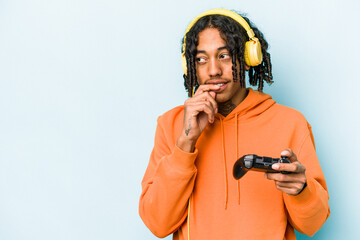 Young African American man playing with a video game controller isolated on blue background relaxed thinking about something looking at a copy space.