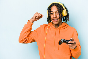 Young African American man playing with a video game controller isolated on blue background raising fist after a victory, winner concept.