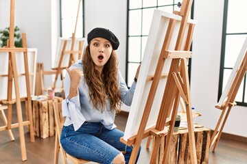 Young hispanic artist woman painting on canvas at art studio surprised pointing with hand finger to the side, open mouth amazed expression.