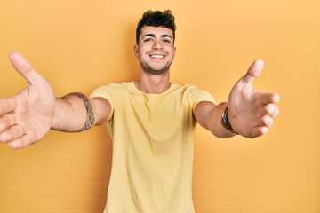 Young hispanic man wearing casual t shirt looking at the camera smiling with open arms for hug. cheerful expression embracing happiness.