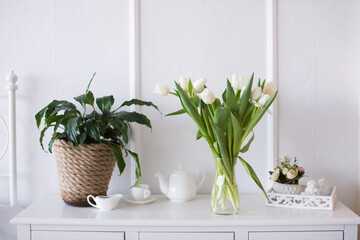 White tulips and a green indoor flower stand on a white table next to tea dishes