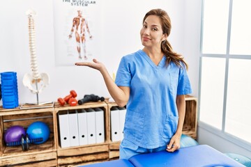 Middle age hispanic physiotherapist woman working at pain recovery clinic smiling cheerful presenting and pointing with palm of hand looking at the camera.