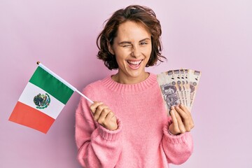 Young brunette woman holding mexico flag and mexican pesos banknotes winking looking at the camera...