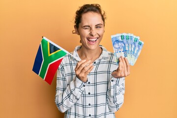 Young brunette woman holding south africa flag and rand banknotes winking looking at the camera...