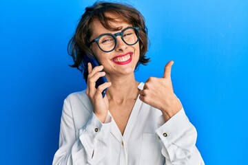 Young brunette woman having conversation talking on the smartphone smiling happy and positive, thumb up doing excellent and approval sign