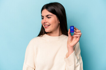 Young caucasian woman holding a batterie isolated on blue background looks aside smiling, cheerful and pleasant.