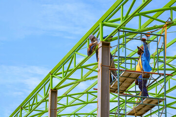 Fototapeta premium Asian foreman and construction worker on scaffolding are welding metal roof structure of industrial building against blue sky in perspective side view