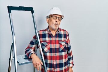 Handsome mature handyman close to construction stairs wearing hardhat making fish face with lips, crazy and comical gesture. funny expression.